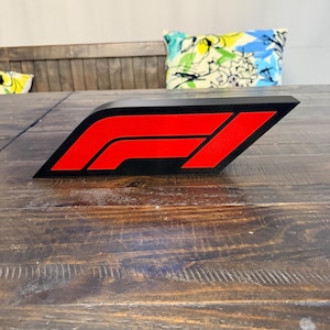 May include: A black and red Formula 1 logo, a stylized geometric design, is displayed on a wooden surface. The logo features a red 'F' and a red stripe against a black background. The design is angled, creating a dynamic visual.