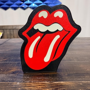 May include: A black, red, and white Rolling Stones tongue and lips logo. The iconic logo is a stylized red tongue and lips with white teeth and a black outline. The item is displayed on a wooden surface.