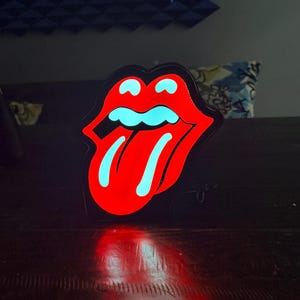 May include: A red and black illuminated sign in the shape of a mouth with a protruding tongue. The interior of the mouth is a bright turquoise color, and the sign is lit up, casting a red glow on the surface below.