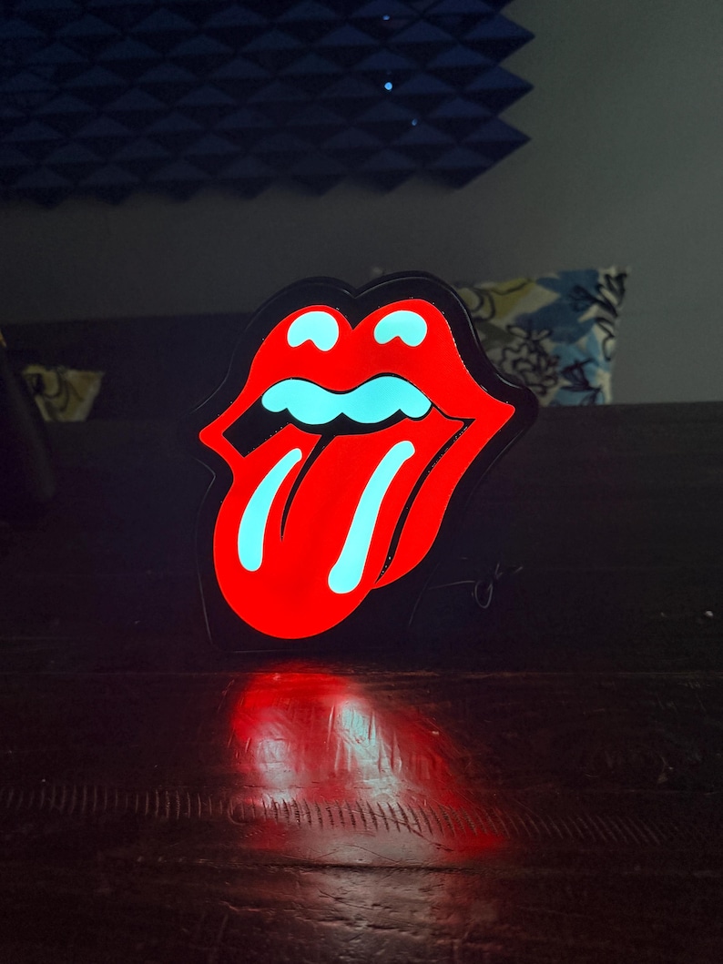 May include: A red and black illuminated sign in the shape of a mouth with a protruding tongue. The interior of the mouth is a bright turquoise color, and the sign is lit up, casting a red glow on the surface below.