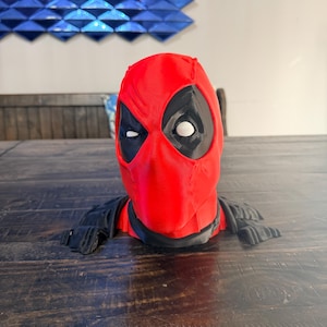 Könnte beinhalten: Eine rot-schwarze Deadpool-Büste mit der ikonischen Maske der Figur mit weißen Augen und einer schwarzen Augenmaske. Die Büste enthält eine schwarze Schulterpanzerung. Der Artikel wird auf einer Holzoberfläche präsentiert.