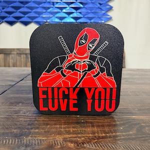 Può includere: Quadrato nero con una grafica rossa di Deadpool che forma un cuore con le mani. Il testo rosso "FUCK YOU" è stampato sotto. Lo sfondo è un motivo geometrico blu.