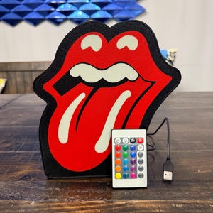 May include: A red and black Rolling Stones tongue and lips logo LED light with a remote control. The light has a black border and a white tongue. The remote has multiple color options and a USB cable.