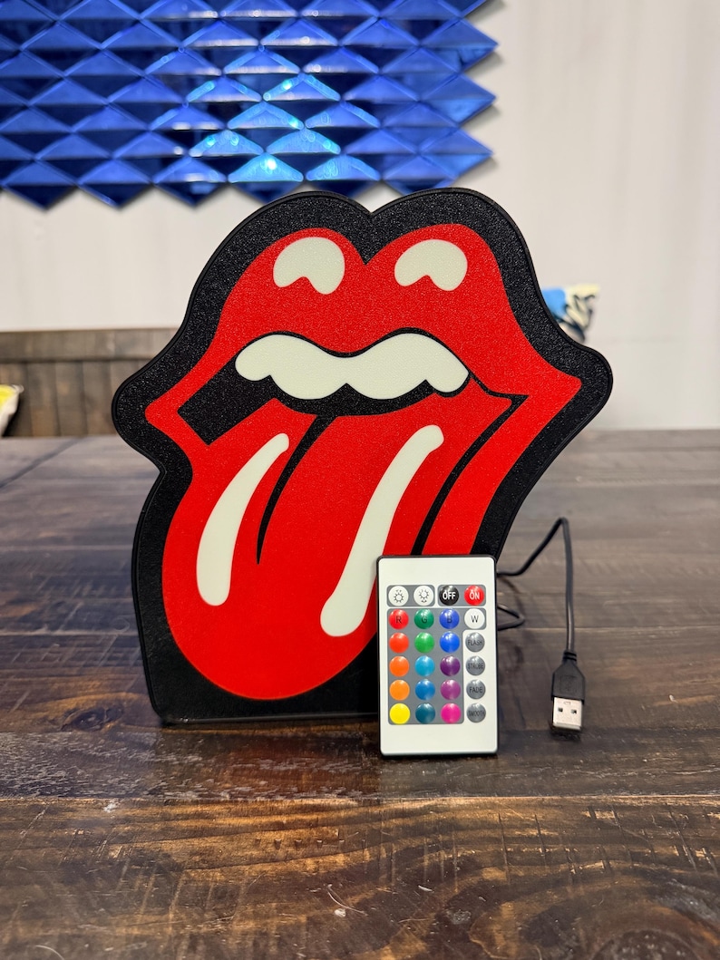 May include: A red and black Rolling Stones tongue and lips logo LED light with a remote control. The light has a black border and a white tongue. The remote has multiple color options and a USB cable.