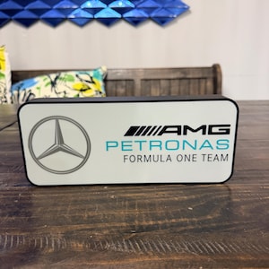 May include: A rectangular white and black sign featuring the AMG Petronas Formula One Team logo. The sign has the Mercedes-Benz logo on the left and the AMG logo with the text "PETRONAS FORMULA ONE TEAM" in teal.