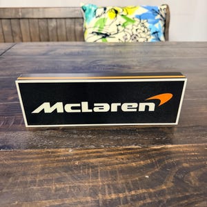 May include: A black rectangular box with the word "McLaren" in white lettering and an orange swoosh logo. The box has an orange stripe along the top edge. The box is on a dark wooden surface.