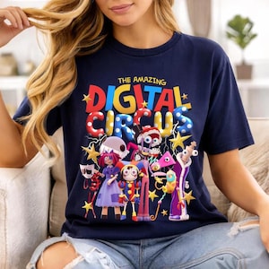 May include: Navy blue t-shirt featuring the text "THE AMAZING DIGITAL CIRCUS" in colorful, 3D bubble letters. The graphic includes cartoon characters, stars, and other circus-themed elements. The shirt is made of a soft material.