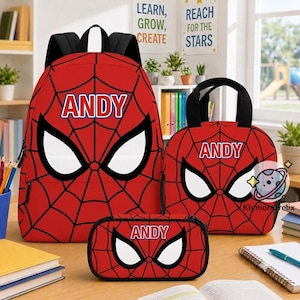 Op de afbeelding: Een rode rugzak, een lunchtas en een etui, allemaal voorzien van een zwart spinnenwebpatroon, witte oogvormen en de naam ANDY in witte letters. Elk item heeft zwarte riemen of handvatten en een bijpassend rood en zwart kleurenschema.