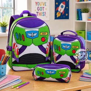 Op de afbeelding: Een set bestaande uit een rugzak, lunchtas en etui met een Buzz Lightyear ruimtepakontwerp in paars, groen en wit. Elk item toont de naam Andy op een geel rechthoekig label, samen met rode, blauwe en groene details van een bedieningspaneel.