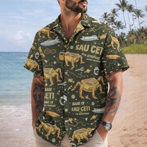 May include: Dark green button-up shirt with a repeating pattern of yellow space-themed creatures, spacecraft, and tools. The shirt has text that reads "SAU CET" and "NOW ASTROPHAGE". The short-sleeved shirt is worn on a beach.