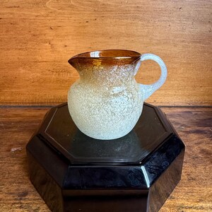 May include: A small, frosted glass pitcher with a brown rim and handle. The pitcher has a rounded body and a spout for pouring. It sits on a black, octagonal base against a wooden background.