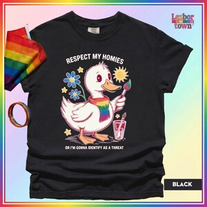 Puede incluir: Camiseta negra con un pato de dibujos animados con una bufanda arco&iacute;ris y un pincel. El texto "Respect My Homies" y "Or I'm Gonna Identify As A Threat" est&aacute; impreso. Una bandera y una pulsera arco&iacute;ris son visibles.