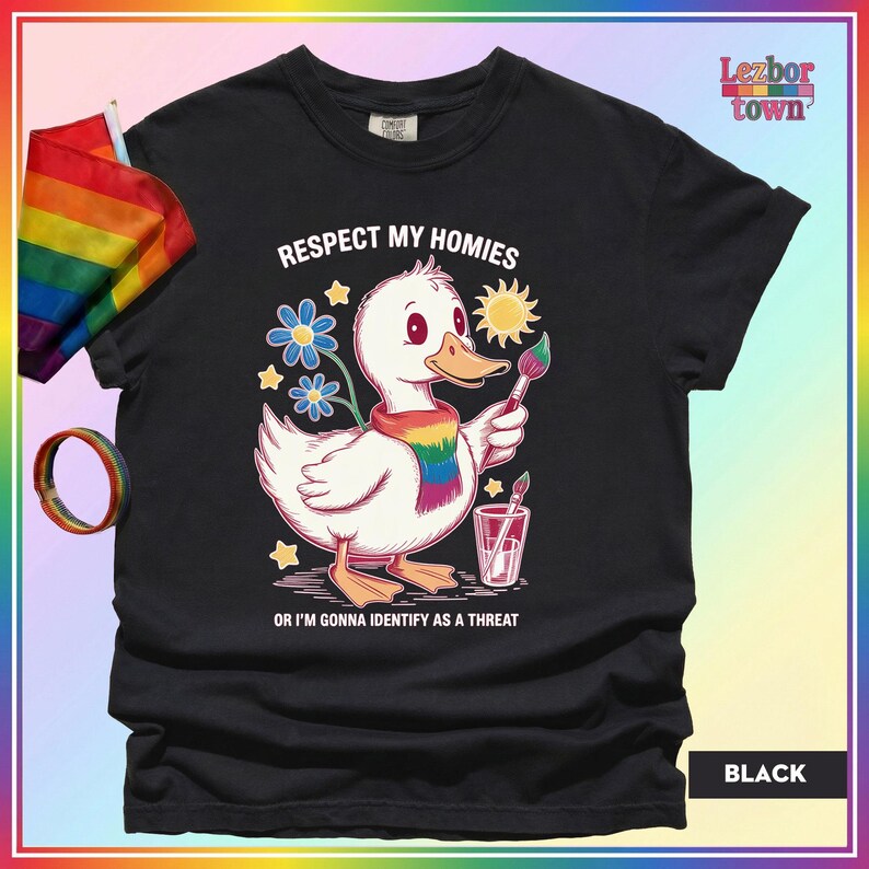 Puede incluir: Camiseta negra con un pato de dibujos animados con una bufanda arco&iacute;ris y un pincel. El texto "Respect My Homies" y "Or I'm Gonna Identify As A Threat" est&aacute; impreso. Una bandera y una pulsera arco&iacute;ris son visibles.
