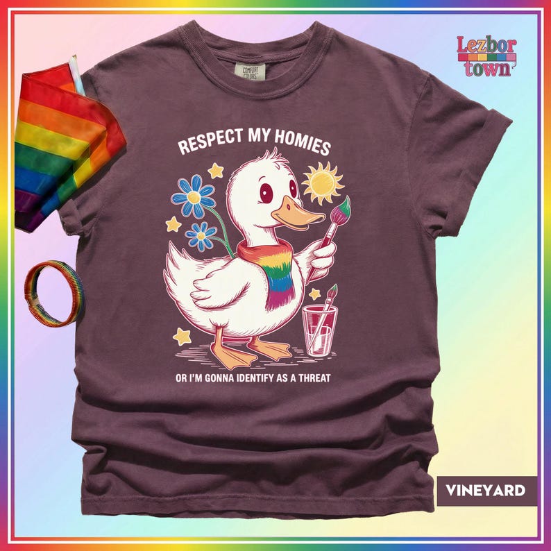 Puede incluir: Camiseta granate con un pato de dibujos animados con una bufanda arco&iacute;ris y un pincel. El texto "Respect My Homies" y "Or I'm Gonna Identify As A Threat" est&aacute; impreso. Una bandera y una pulsera arco&iacute;ris son visibles.