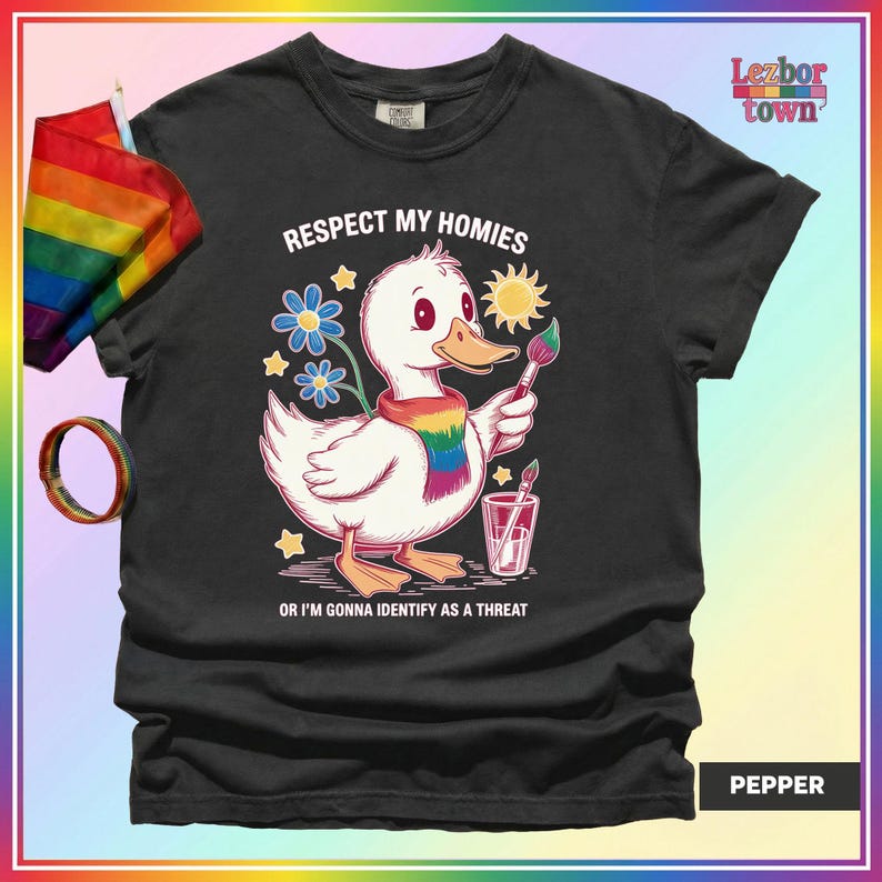 Puede incluir: Camiseta negra con un dibujo de un pato blanco con una bufanda arco&iacute;ris, sosteniendo un pincel y el texto "RESPECT MY HOMIES OR I'M GONNA IDENTIFY AS A THREAT". Se ven una bandera y una pulsera arco&iacute;ris.