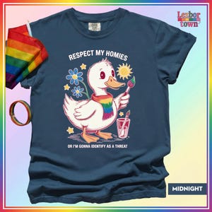 Puede incluir: Camiseta azul medianoche con un dibujo de un pato blanco con una bufanda arco&iacute;ris y un pincel. El texto dice "Respect My Homies" y "Or I'm Gonna Identify As A Threat". Una bandera y una pulsera arco&iacute;ris son visibles.