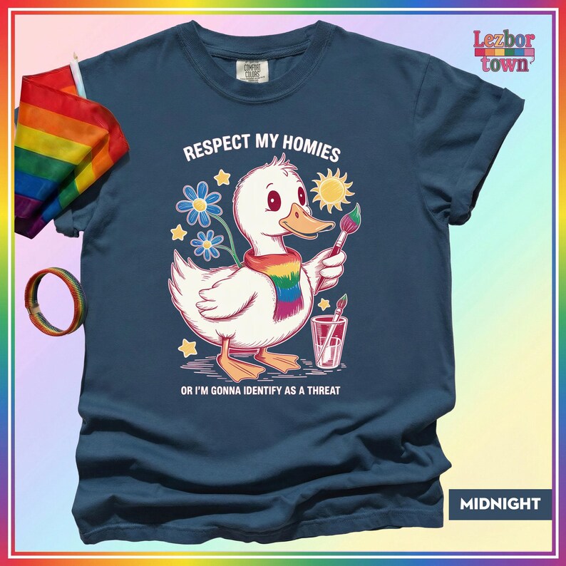 Puede incluir: Camiseta azul medianoche con un dibujo de un pato blanco con una bufanda arco&iacute;ris y un pincel. El texto dice "Respect My Homies" y "Or I'm Gonna Identify As A Threat". Una bandera y una pulsera arco&iacute;ris son visibles.