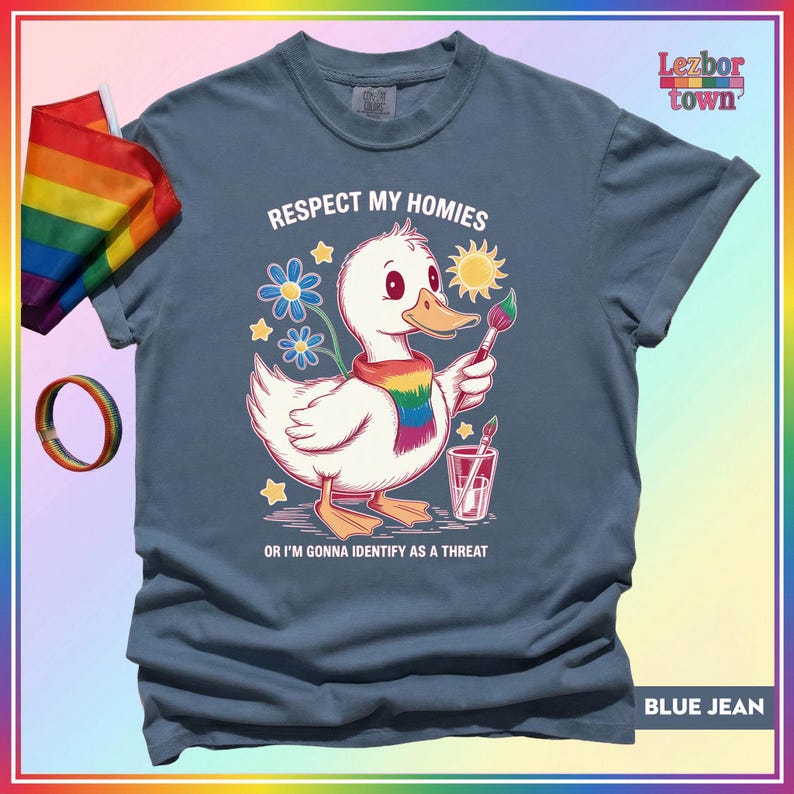 Puede incluir: Camiseta azul vaquero con un pato de dibujos animados que lleva una bufanda arco&iacute;ris y sostiene un pincel. El texto de la camiseta dice: "Respect my homies or I'm gonna identify as a threat". Tambi&eacute;n se ven una bandera y una pulsera arco&iacute;ris.