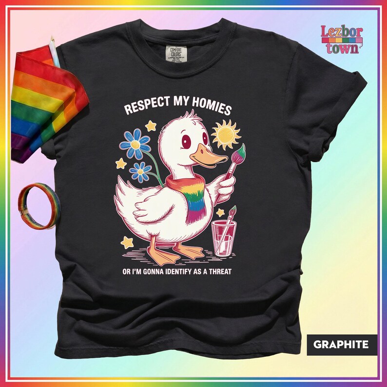 Puede incluir: Camiseta de color grafito con un pato de dibujos animados con una bufanda arco&iacute;ris y un pincel. El texto "Respect My Homies Or I'm Gonna Identify As A Threat" est&aacute; impreso. Una bandera y una pulsera arco&iacute;ris son visibles.