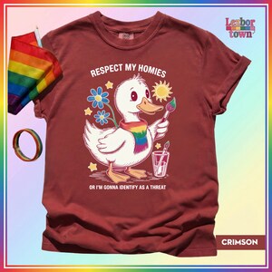 Puede incluir: Camiseta color carmes&iacute; con un dibujo de un pato blanco con una bufanda arco&iacute;ris y un pincel. El texto dice "Respect My Homies" y "Or I'm Gonna Identify As A Threat". Tambi&eacute;n se ven una bandera y una pulsera arco&iacute;ris.