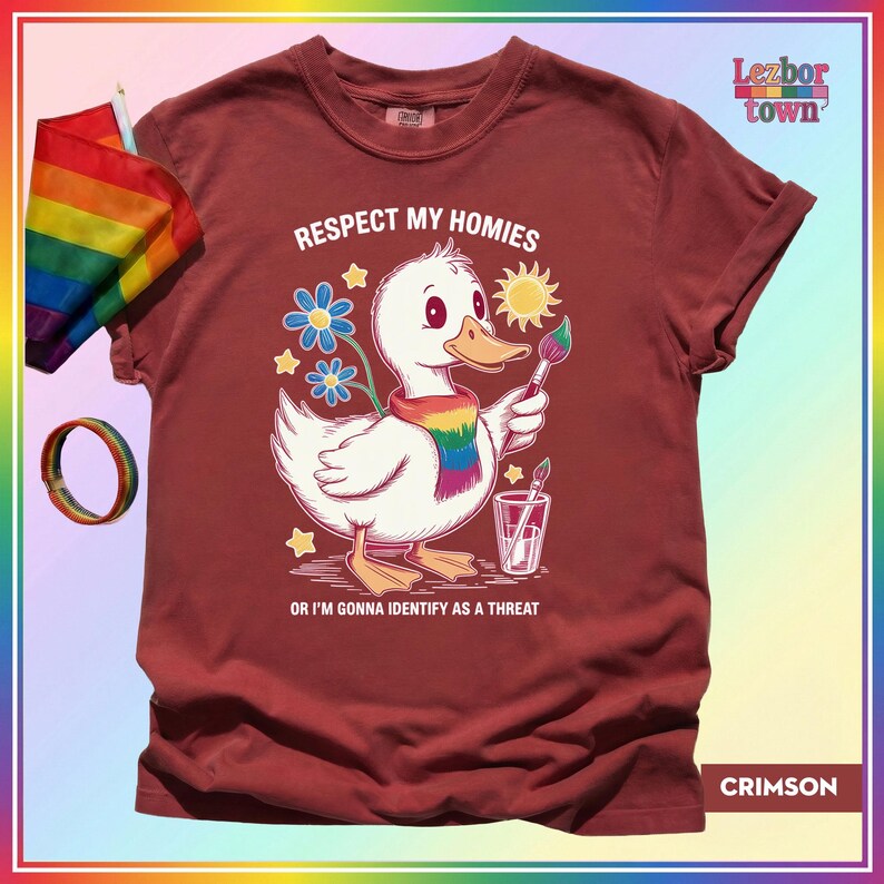 Puede incluir: Camiseta color carmes&iacute; con un dibujo de un pato blanco con una bufanda arco&iacute;ris y un pincel. El texto dice "Respect My Homies" y "Or I'm Gonna Identify As A Threat". Tambi&eacute;n se ven una bandera y una pulsera arco&iacute;ris.