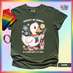 Puede incluir: Camiseta verde musgo con un dibujo de un pato blanco con una bufanda arco&iacute;ris y un pincel. El texto dice "Respect My Homies" y "Or I'm Gonna Identify As A Threat". Una bandera y una pulsera arco&iacute;ris son visibles.