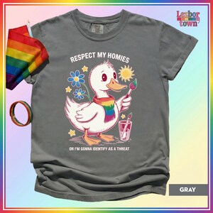 Puede incluir: Camiseta gris con un pato de dibujos animados con una bufanda arco&iacute;ris y un pincel. El texto "Respect My Homies" y "Or I'm Gonna Identify As A Threat" est&aacute; impreso. Una bandera y una pulsera arco&iacute;ris son visibles.