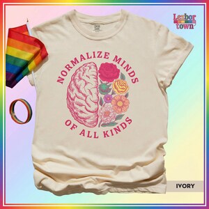 Puede incluir: Camiseta de color marfil con el texto "Normalize Minds of All Kinds" que rodea un cerebro y un diseño floral. Una bandera y una pulsera arcoíris son visibles a la izquierda. La camiseta es de la marca Comfort Colors.