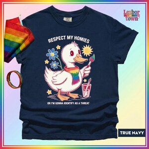 Puede incluir: Camiseta azul marino con un pato de dibujos animados con una bufanda arco&iacute;ris y un pincel. El texto de la camiseta dice: "Respect My Homies or I'm Gonna Identify as a Threat." Tambi&eacute;n se ven una bandera y una pulsera arco&iacute;ris.