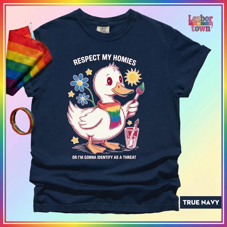 Puede incluir: Camiseta azul marino con un pato de dibujos animados con una bufanda arco&iacute;ris y un pincel. El texto de la camiseta dice: "Respect My Homies or I'm Gonna Identify as a Threat." Tambi&eacute;n se ven una bandera y una pulsera arco&iacute;ris.