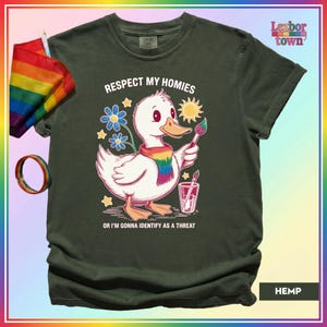 Puede incluir: Camiseta verde oscuro con un pato de dibujos animados con una bufanda arco&iacute;ris y un pincel. El texto en la camiseta dice "Respect My Homies" y "Or I'm Gonna Identify As A Threat". Tambi&eacute;n se ven una bandera y una pulsera arco&iacute;ris.