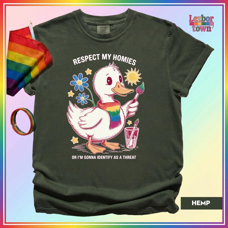 Puede incluir: Camiseta verde oscuro con un pato de dibujos animados con una bufanda arco&iacute;ris y un pincel. El texto en la camiseta dice "Respect My Homies" y "Or I'm Gonna Identify As A Threat". Tambi&eacute;n se ven una bandera y una pulsera arco&iacute;ris.