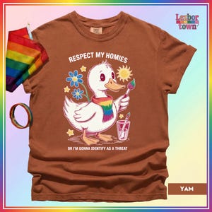 Puede incluir: Camiseta marr&oacute;n con un dibujo de un pato blanco con una bufanda arco&iacute;ris y un pincel. El texto de la camiseta dice: "Respect My Homies Or I'm Gonna Identify As A Threat." Una bandera y una pulsera arco&iacute;ris son visibles.
