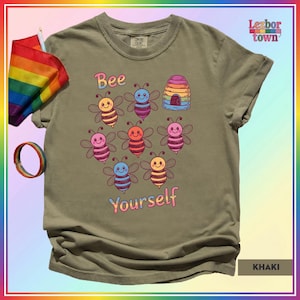 Può includere: T-shirt color cachi con la scritta "Bee Yourself" e una grafica di api e un alveare. Le api sono di vari colori. Una bandiera e un braccialetto arcobaleno sono nell'angolo in alto a sinistra.