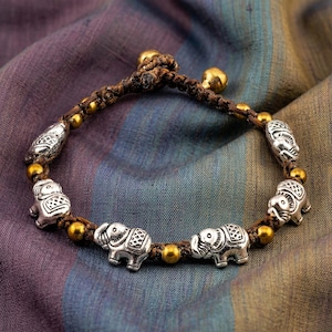 May include: A handmade bracelet featuring silver elephant charms and gold beads strung on a brown cord. The bracelet has a woven design and is accented with gold-colored bells. The background is a multicolored fabric.