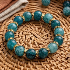 May include: A teal beaded bracelet with gold-colored spacers. The bracelet features large, round beads with a marbled texture. The gold spacers are adorned with small, clear crystals. The bracelet is displayed on a woven surface.