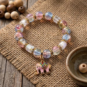 May include: A beaded bracelet with a pink elephant charm. The bracelet features iridescent, multi-colored beads and gold accents. The elephant charm is pink with colorful gem details. The bracelet is displayed on a burlap surface.