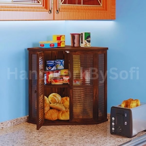 May include: A dark wooden corner cabinet with glass-paneled doors, filled with canned goods, bread, and other food items. The cabinet sits on a countertop next to a toaster with two slices of toast. The text "Handmade By Sofi" is visible.