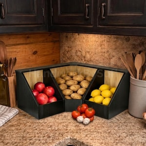May include: Three-section wooden produce storage unit with black exterior and light wood interior. Each section holds different fruits and vegetables: red apples, potatoes, and lemons. Tomatoes and garlic are in front.