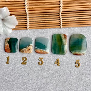 May include: Five polished, rectangular cabochons with rounded corners, featuring swirling patterns of teal, green, and tan. Each stone is labeled with a gold-colored number from one to five, arranged in a horizontal row on a textured surface.