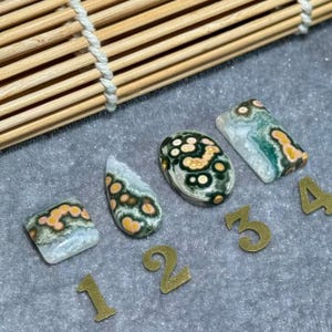 Può includere: Quattro pietre preziose lucide con motivi e colori unici. Le pietre sono numerate da 1 a 4 con numeri metallici color oro. Le pietre preziose hanno un mix di colori verdi, arancioni e bianchi. Sullo sfondo una persiana di bambù in legno.