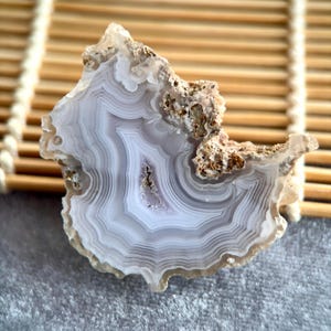 May include: A polished agate stone slice with concentric bands of gray and white. The natural stone has a rough, textured edge with areas of brown and tan. The stone is displayed on a light brown bamboo mat.