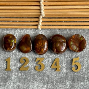 May include: Five polished, oval-shaped gemstones in shades of brown and amber, each with unique internal patterns. The gemstones are numbered 1 through 5 with gold-colored numerals. The background is a gray fabric.