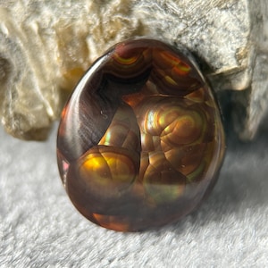 May include: A polished, teardrop-shaped fire agate gemstone featuring deep brown and black tones with iridescent, glowing orange and gold orb-like inclusions that shimmer across the smooth, rounded surface of the translucent stone.