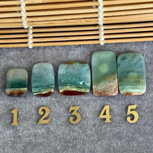 May include: Five rectangular jade stones with rounded corners, each displaying a unique blend of teal, brown, and white hues. The stones are arranged in a row, numbered 1 through 5 with gold-colored numerals. A bamboo mat is in the background.