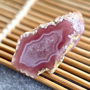 May include: A polished agate stone with concentric bands of pink and white, displaying a central crystalline cavity. The outer edges have a rough, textured appearance with hints of yellow and green.