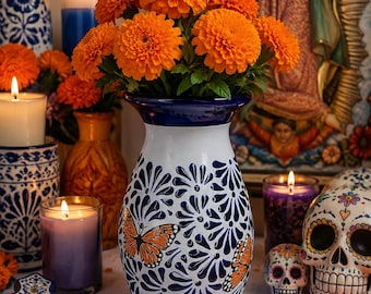 May include: A white ceramic vase with a dark blue rim and a hand-painted pattern of dark blue floral motifs and orange monarch butterflies. The vase is filled with a bouquet of bright orange marigold flowers, creating a vibrant display.
