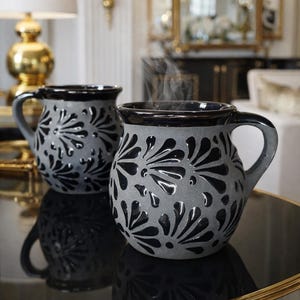Puede incluir: Dos tazas de cerámica gris con diseños florales negros y asas. El vapor se eleva de las bebidas calientes en su interior. Las tazas están sobre una superficie negra reflectante.