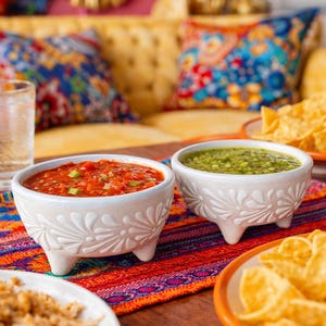 May include: A colourful image of a Mexican feast. Two white bowls with salsa and guacamole sit on a table with tortilla chips, shredded meat, and lime wedges. A patterned runner adds to the vibrant scene.