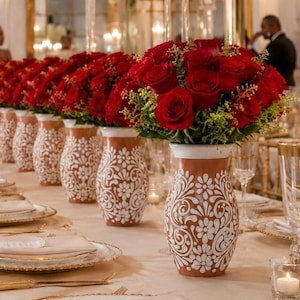 Könnte beinhalten: Eine Reihe von Terrakotta-Vasen mit komplizierten weißen Blumenmustern, die jeweils ein üppiges Arrangement aus tiefroten Rosen und grünem Blattwerk enthalten und in einer Reihe auf einem formellen Esstisch mit Gedecken stehen.